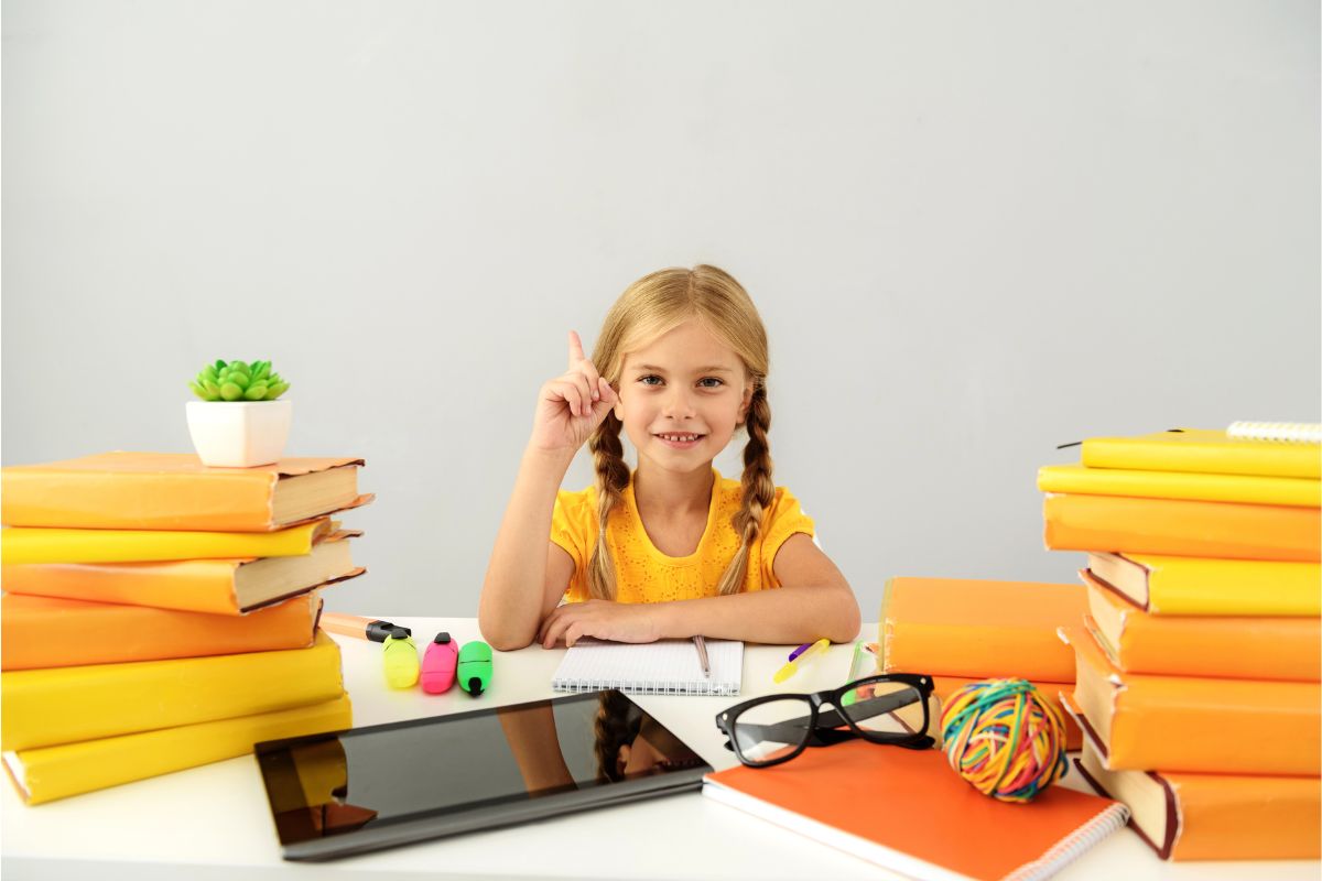 Anak perempuan sedang duduk dengan tumpukan buku di kiri dan kanan, kacamata, dan gadget di atas meja.