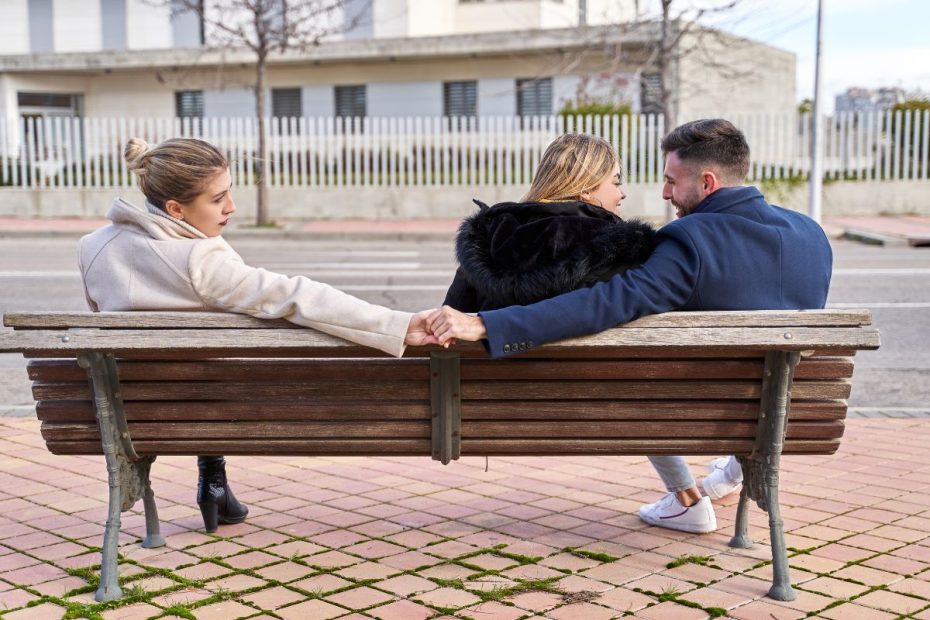 3 orang sedang duduk di bangku taman. Sepasang suami istri duduk berdekatan, tetapi sang suami memegang tangan orang ketiga.