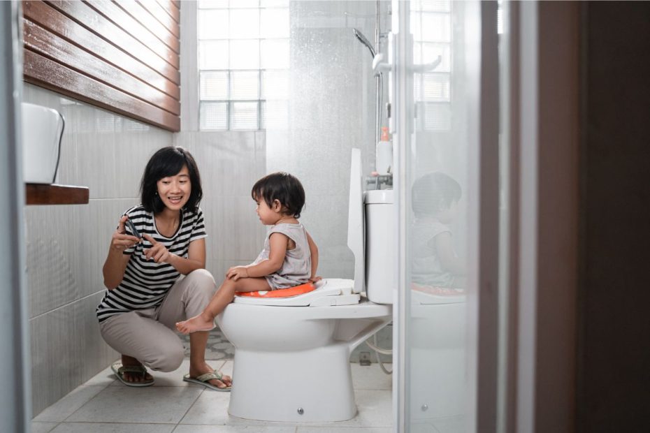 Anak kecil lelaki sedang duduk di kloset bersama sang ibu di depannya sambil diarahkan cara menggunakan toilet.